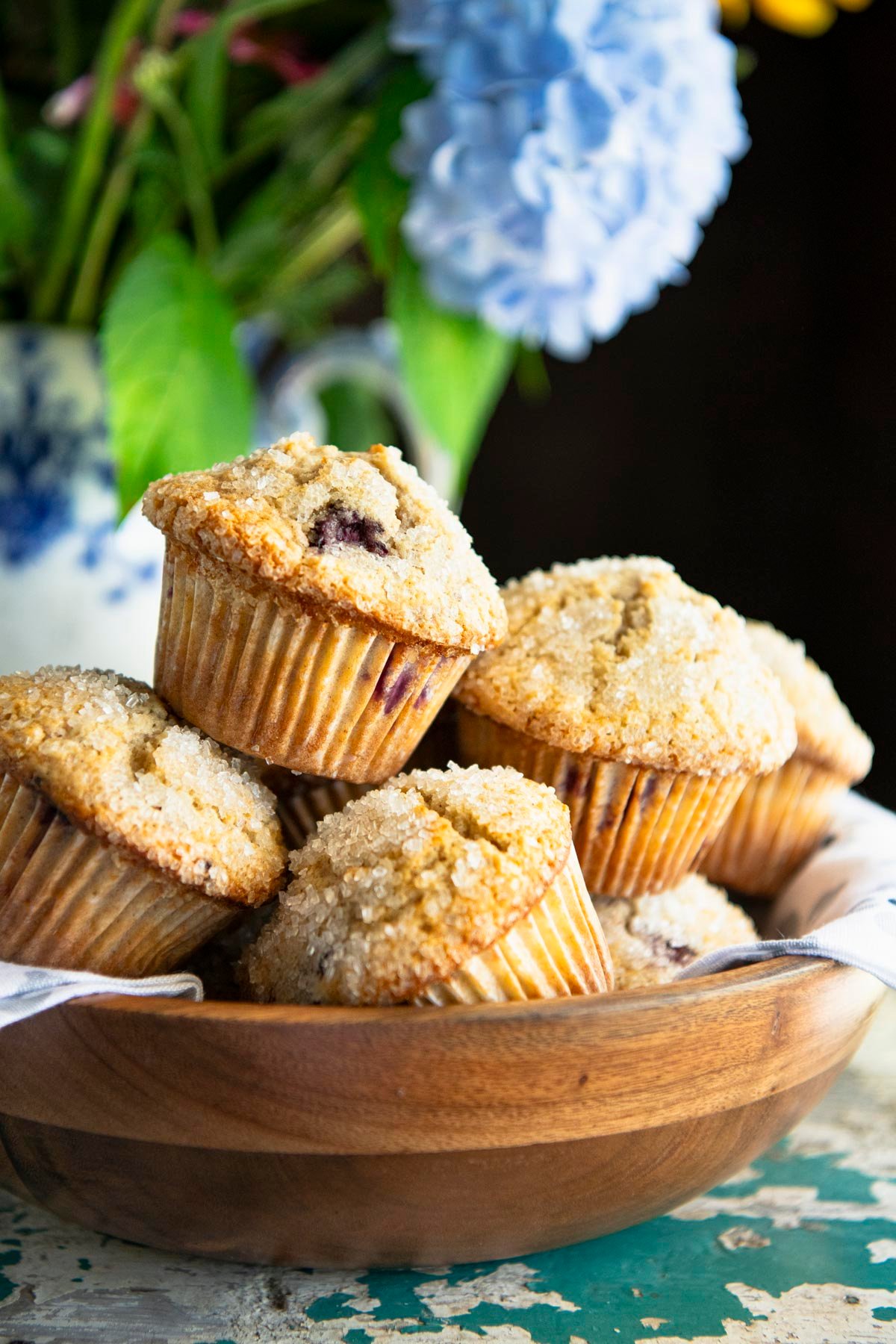 Side shot of blackberry muffins in a wooden bowl with hydrangeas in the background.