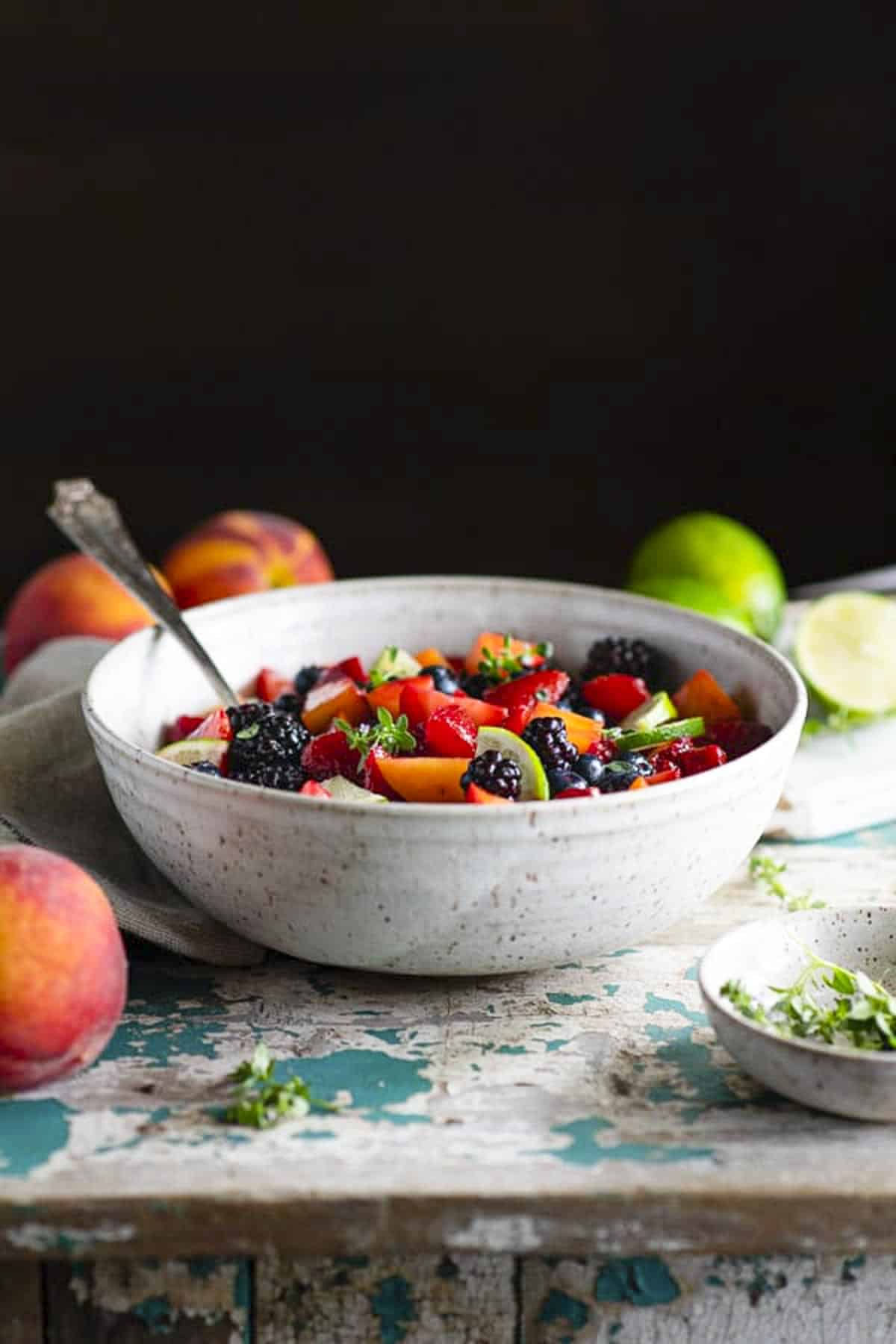 Side shot of an easy fruit salad recipe on a rustic wood table surrounded by peaches and limes.
