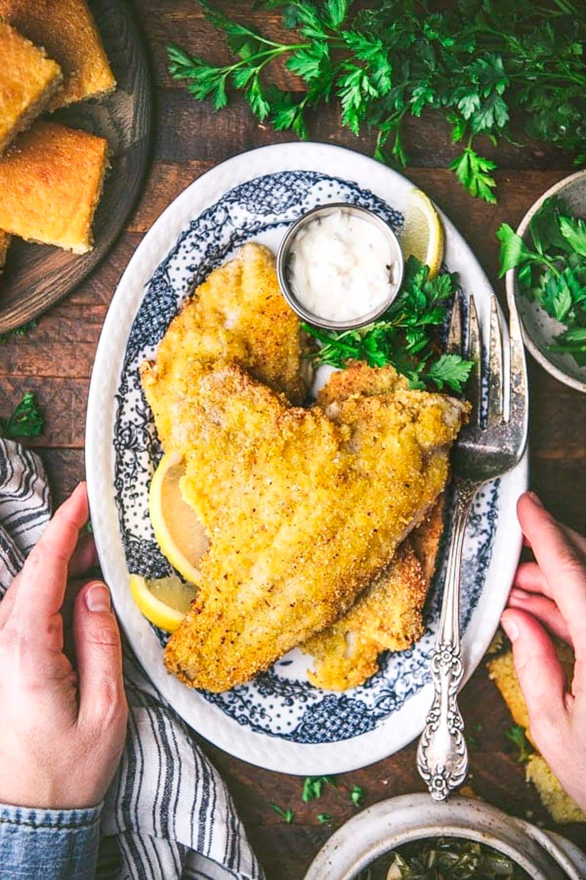 Hands serving a platter of fried catfish on a wooden table.
