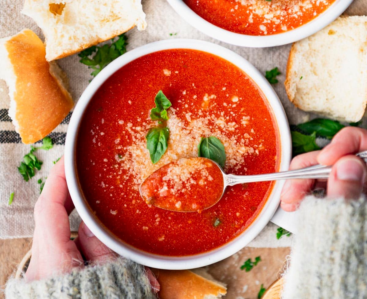 Horizontal overhead image of hands eating a bowl of the best tomato basil soup recipe with a spoon.