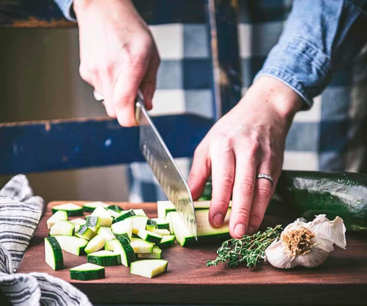 Hands chopping zucchini on a wooden cutting board surrounded by seasonings for zucchini like garlic and thyme.