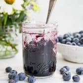 Square side shot of an easy blueberry jam recipe on a white table with fresh flowers in the background.