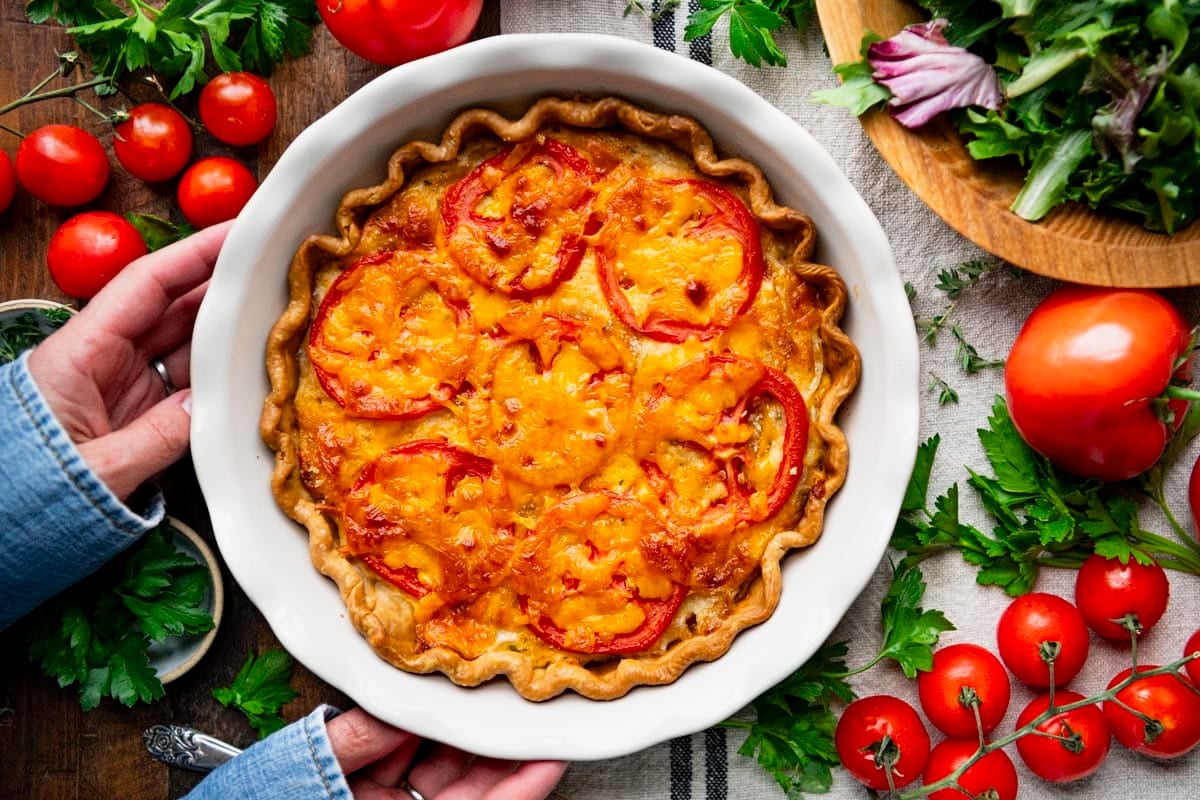 Horizontal overhead image of hands holding a white pie plate with a southern tomato pie in it.