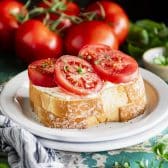 Horizontal image of the best tomato sandwich recipe served on a white plate with fresh basil and tomatoes in the background.