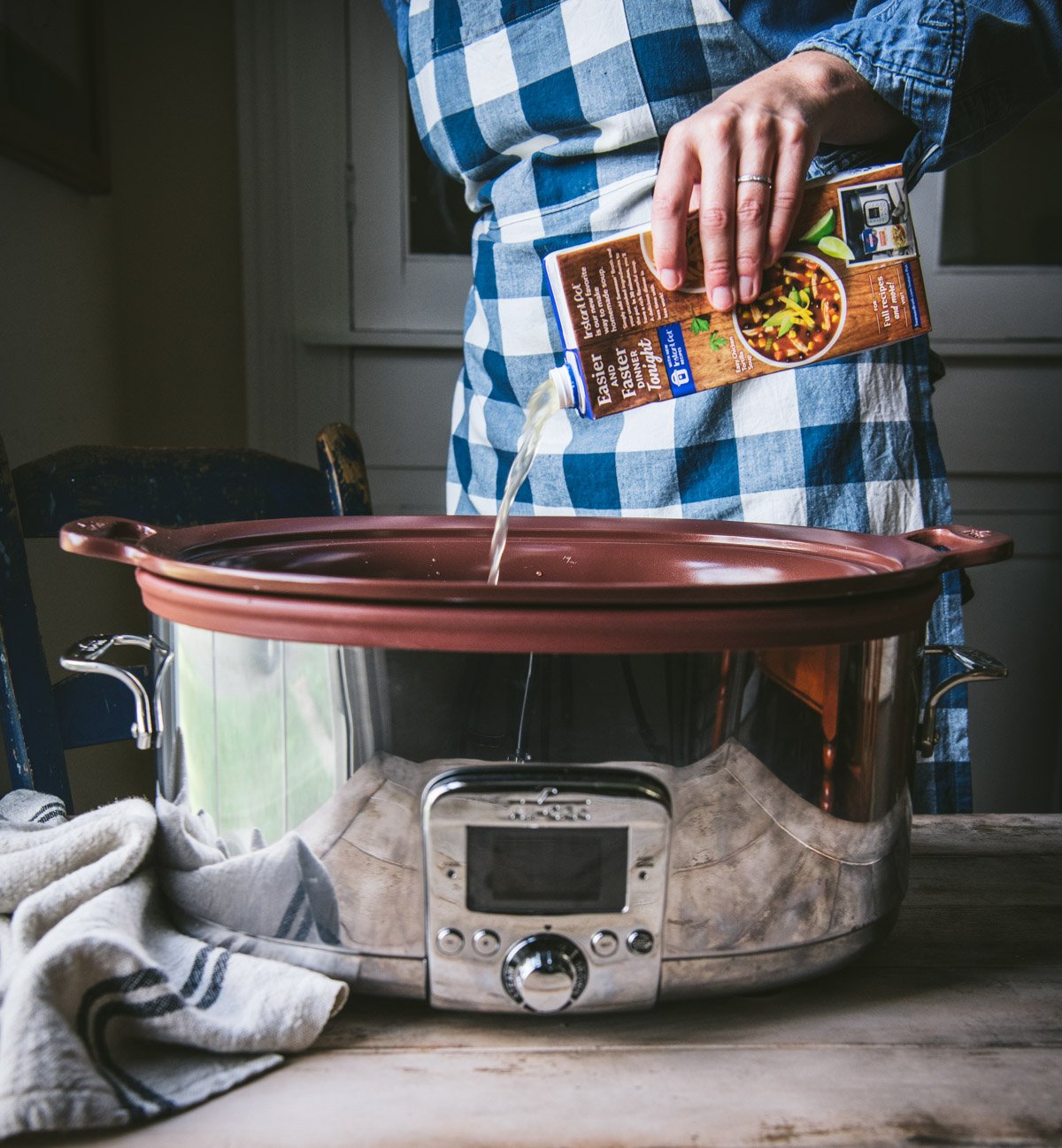 A woman pours a carton of chicken broth into a large crock pot.