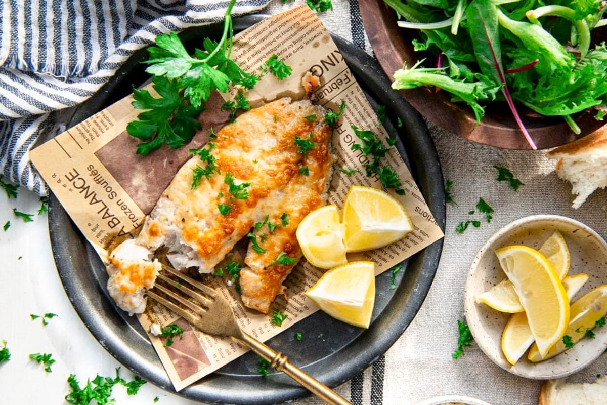 Overhead shot of a plate of garlic parmesan tilapia recipe on a white table with a side salad and lemons.