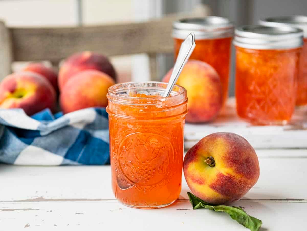 Horizontal side shot of jars of fresh peach jam on a white farmhouse table.