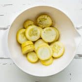 Sliced yellow summer squash in a bowl with olive oil, salt, and pepper.