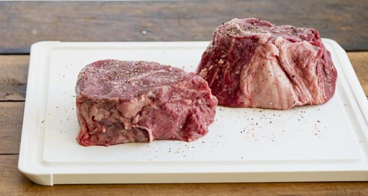 Two filet mignon raw on a cutting board with seasoning.
