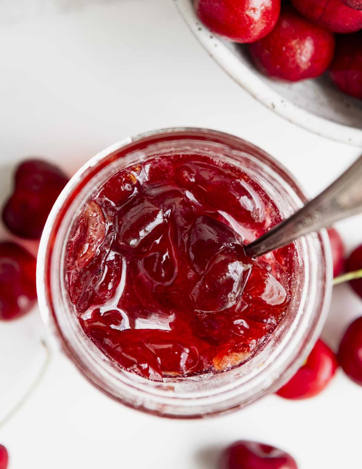 Overhead shot of a spoon in a jar of cherry jam on a white table.
