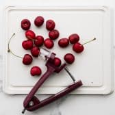 Overhead image of a cherry pitter on white cutting board with fresh cherries.