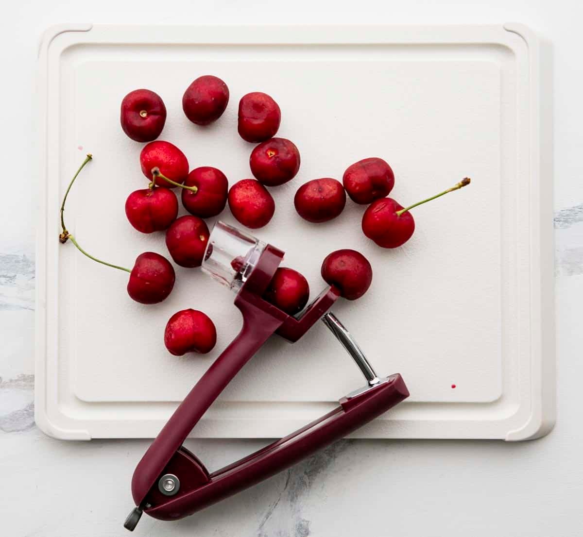 Overhead image of a cherry pitter on white cutting board with fresh cherries.