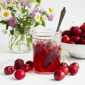 Horizontal side shot of a jar of cherry jam with fresh cherries in a bowl in the background.
