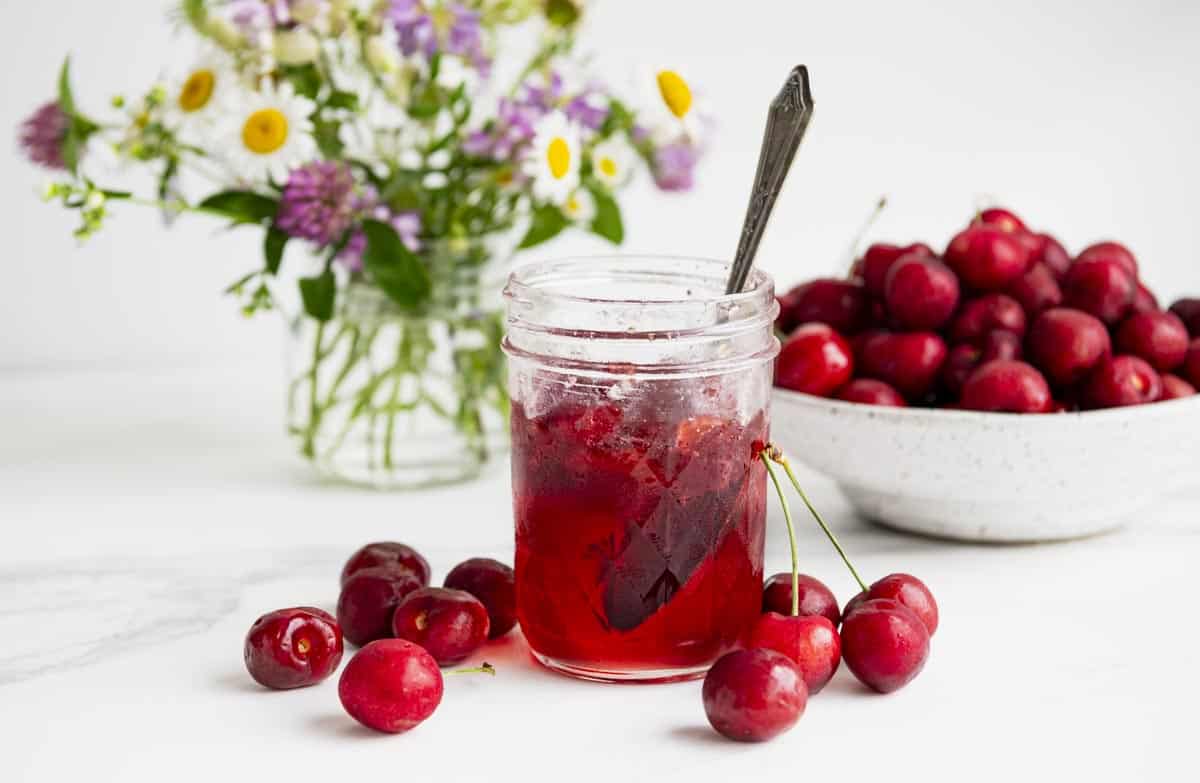 Horizontal side shot of a jar of cherry jam with fresh cherries in a bowl in the background.