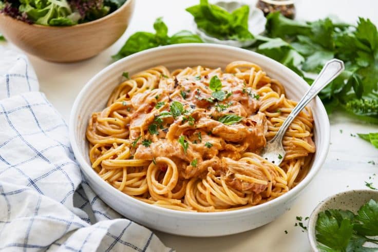 Horizontal image of a white bowl of cream cheese chicken crock pot recipe with pasta and a side salad.
