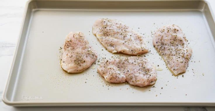 Chicken cutlets on a rimmed baking sheet with Italian seasoning.