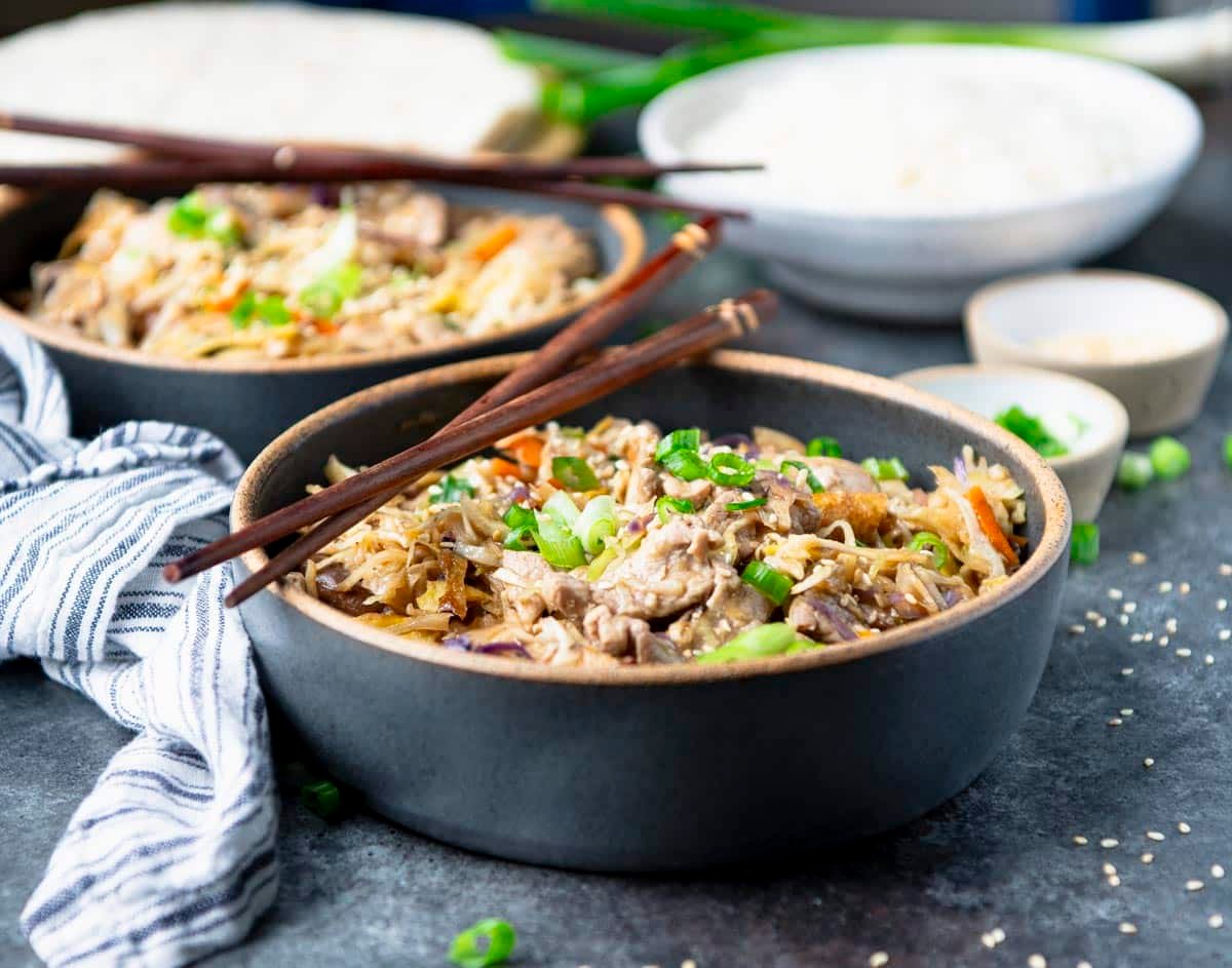 Horizontal image of moo shu pork in bowls with rice and pancakes in the background.