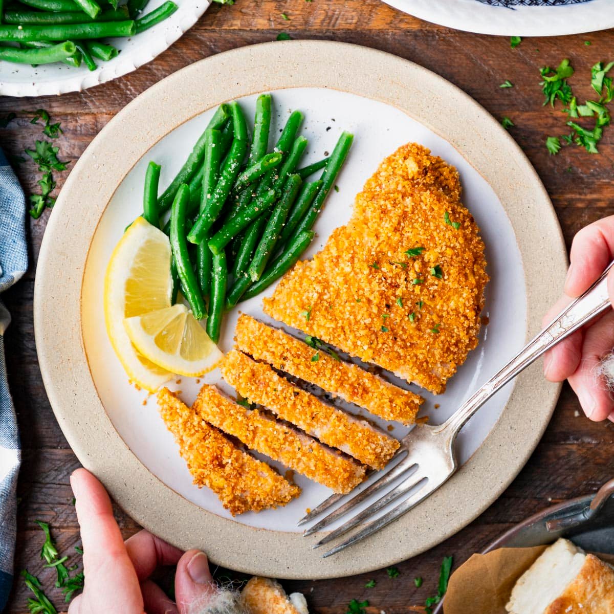 Square overhead image of hands eating baked breaded pork chops on a plate with a fork.
