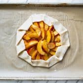 Process shot showing how to make a peach galette on a baking sheet before baking in the oven.