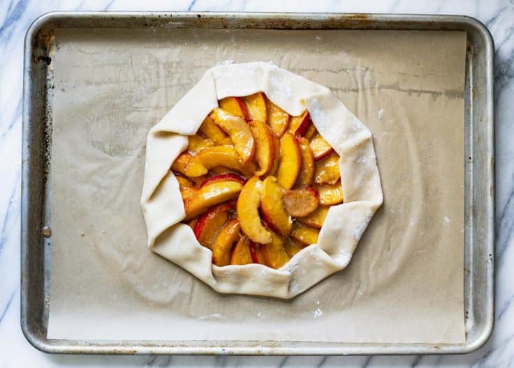 Process shot showing how to make a peach galette on a baking sheet before baking in the oven.