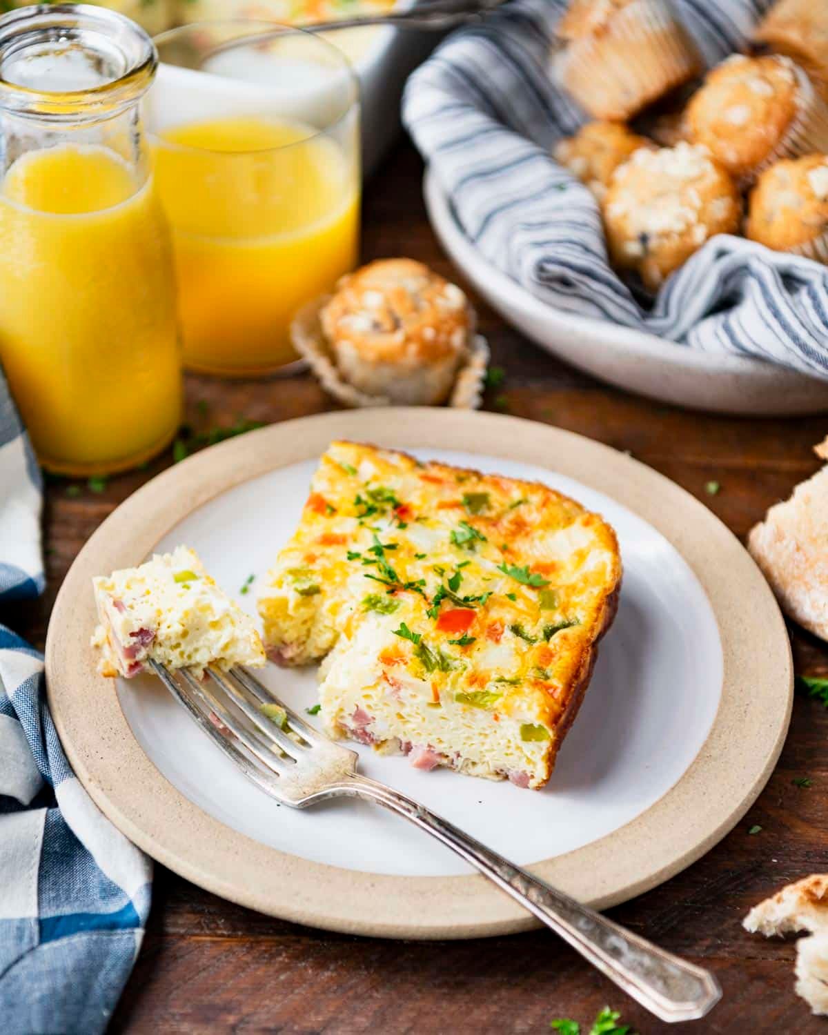 Front shot of a slice of baked Denver omelet on a white plate with orange juice and muffins in the background.