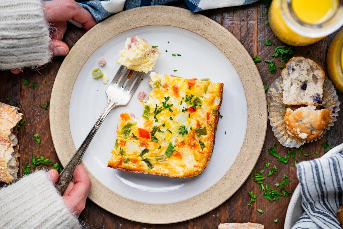 Horizontal overhead shot of hands eating a slice of baked western omelette on a white plate.