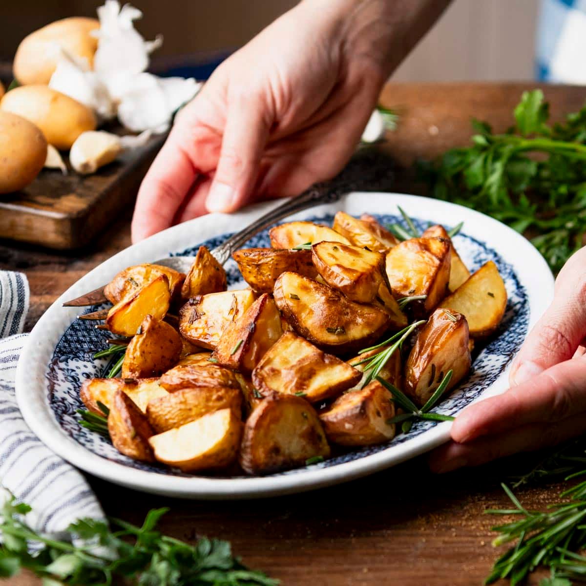 Square side shot of hands serving roasted gold potatoes with rosemary and olive oil.