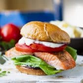 Square side shot of a baked salmon sandwich with lettuce, tomato, and tartar sauce on a brioche bun.