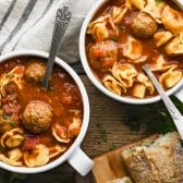 Horizontal overhead shot of two bowls of meatball tortellini soup on a rustic wooden table.