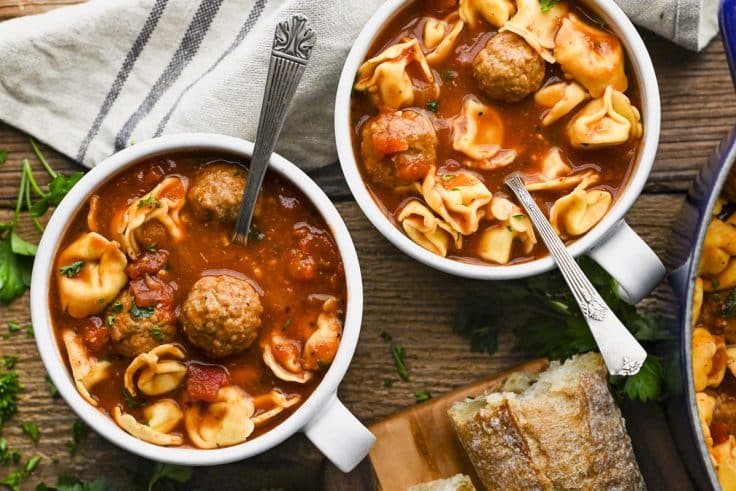 Horizontal overhead shot of two bowls of meatball tortellini soup on a rustic wooden table.